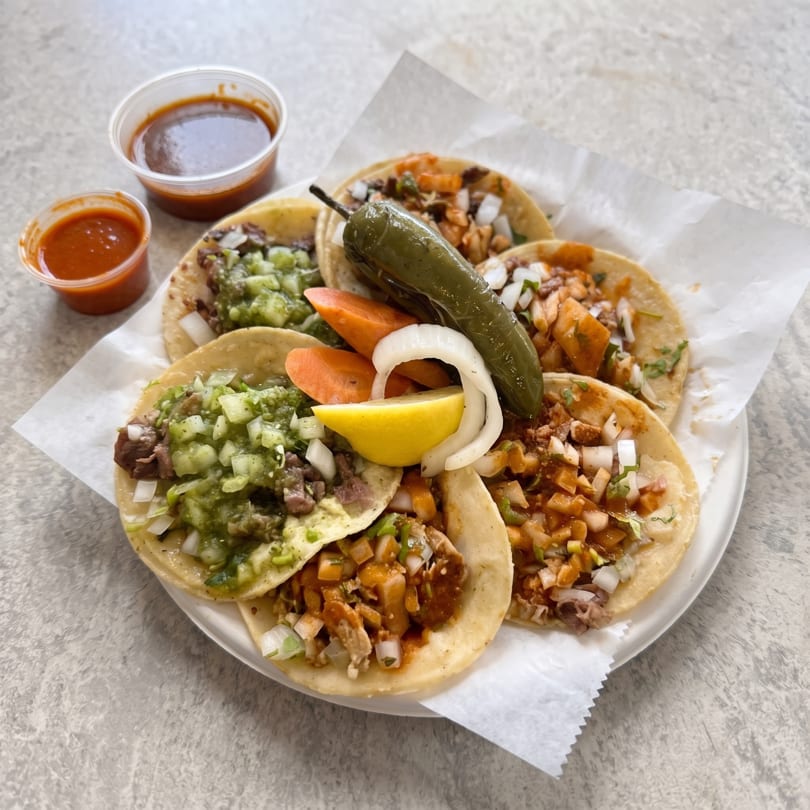A plate of six street tacos with various toppings, salsas, and a jalapeño.