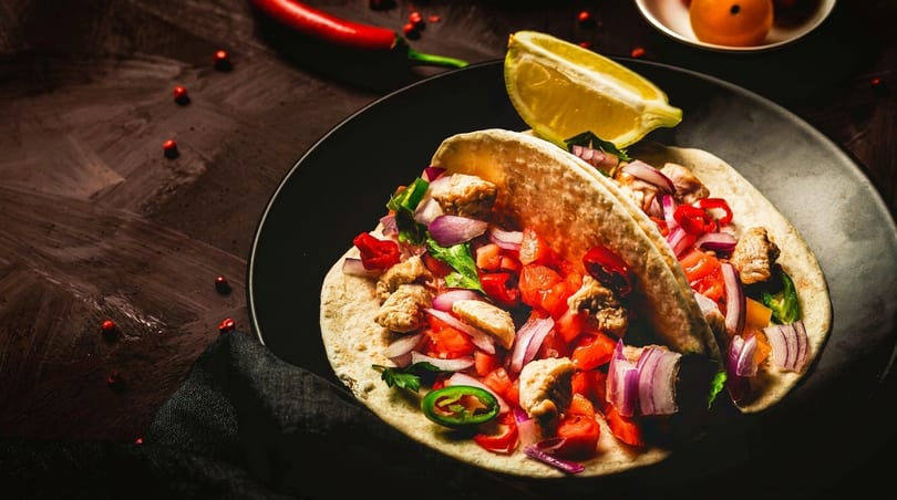 Two chicken tacos with red onion, tomato, and jalapeño, served with a lime wedge.