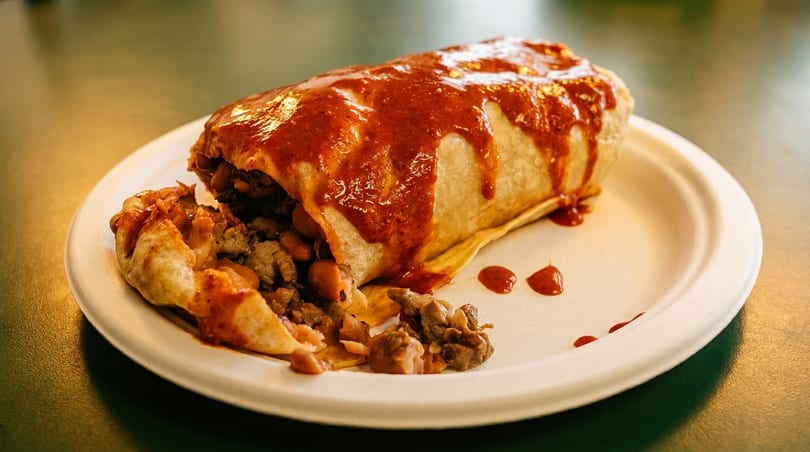 A large burrito, partially unwrapped, covered in red sauce and spilling out meat and beans.