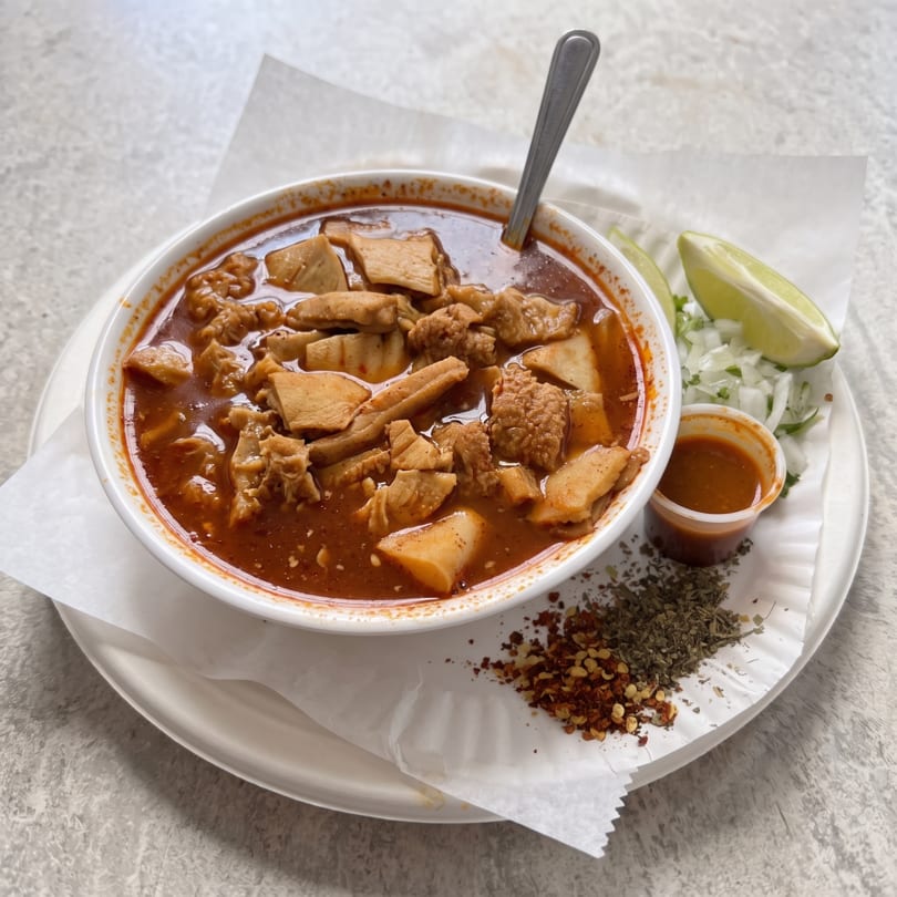 Menudo soup in a white bowl with a spoon, served with lime, onions, chili flakes, and oregano.
