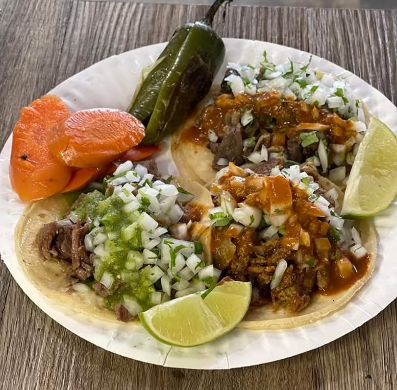 Three street tacos with various toppings, lime wedges, grilled jalapeño, and pickled carrots on a plate.