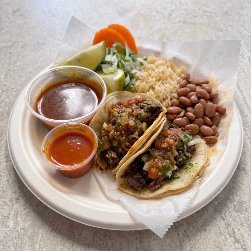 Two beef tacos with salsa, rice, beans, lime, and orange slices on a white plate.