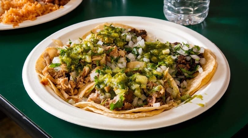 A plate of tacos with various fillings, green salsa, and diced onions on a green table.