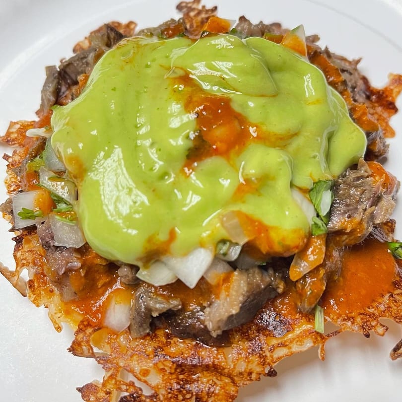 A crispy cheese taco with carne asada, onions, cilantro, red sauce, and green avocado crema.